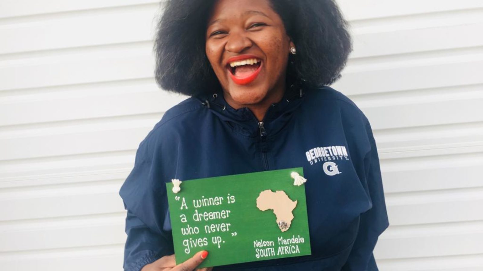 Photo of Maputi Botlhole wearing a Georgetown jacket and smiling, holding a green sign that says &quot;A winner is a dreamer who never gives up. Nelson Mandela, South Africa.&quot;