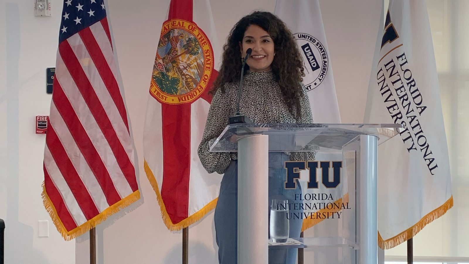 Pierina standing at a glass Florida International University podium in front of an American, Florida, USAID, and FIU flags