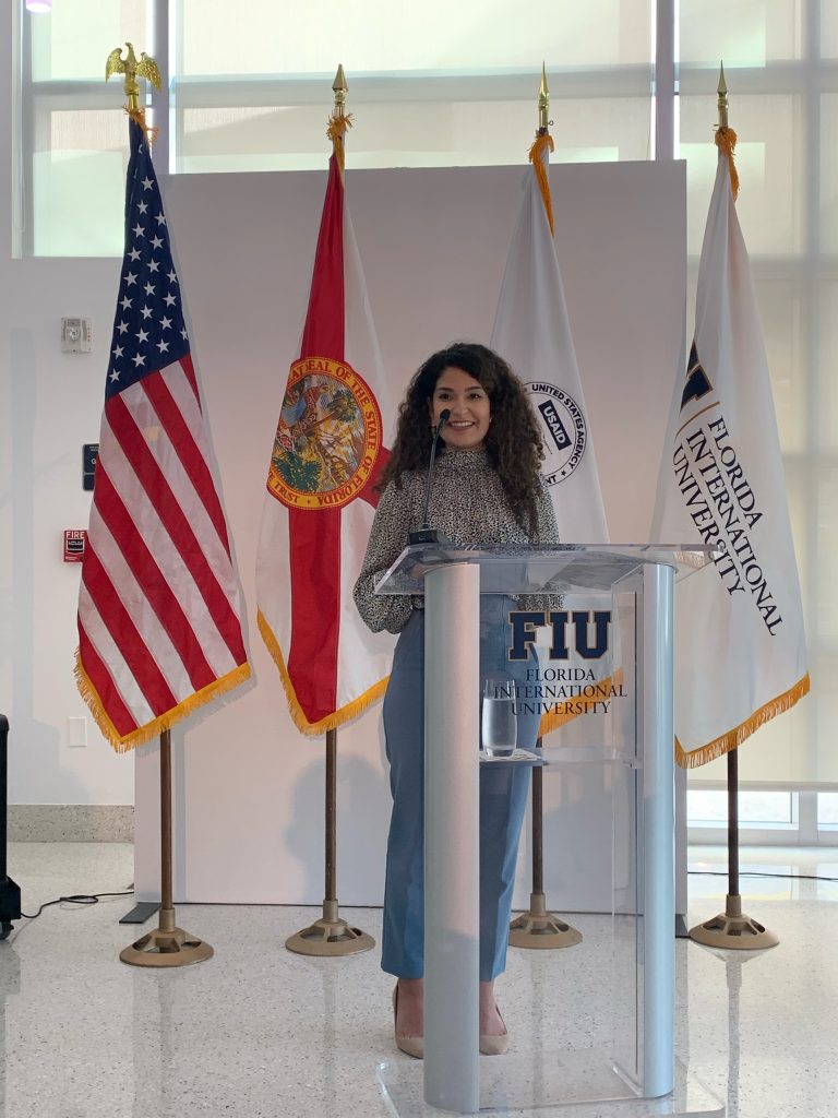 Pierina standing at a glass Florida International University podium in front of an American, Florida, USAID, and FIU flags