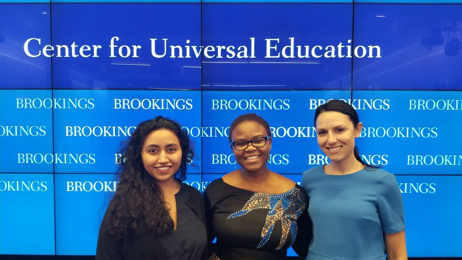 Foluyinka standing between two women in from of a Brookings Center for Universal Education screen