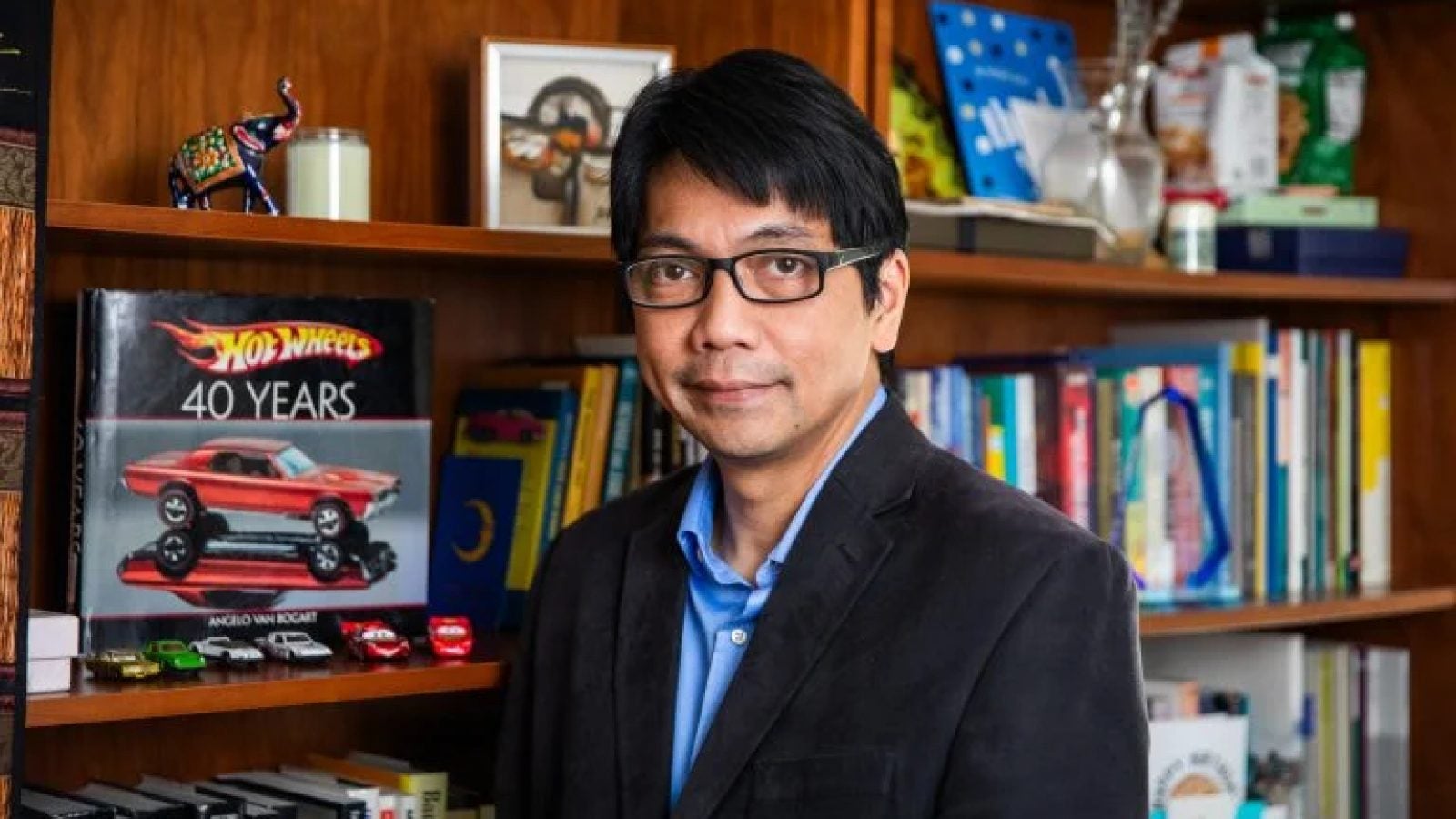 Professor Erwin Tiongson stands in front of a bookshelf displaying Hot Wheels cars